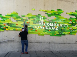 A person and a child interact with a colorful wall display, featuring green rectangular blocks of various sizes, some labeled with text. The display includes words like 'COMMUNITY' and 'COMUNIDAD' prominently. Orange and yellow flowers adorn the display, adding splashes of color.