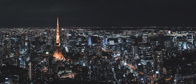 A nighttime cityscape of Tokyo with a brightly illuminated Tokyo Tower standing prominently amidst a sea of city lights and skyscrapers. The lights of the buildings and streets create a shimmering effect against the dark sky.
