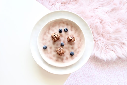 A white plate placed on a textured surface holds energy balls and blueberries. The balls appear to be made of oats or a similar grain. The background includes a pastel pink fluffy fabric, adding a cozy and soft touch to the setting.