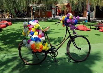 A bicycle is adorned with numerous brightly colored artificial flowers, parked on a green surface in an outdoor area. The setting includes red bean bags scattered around, and a small bar or shop in the background with hanging decorations. Tree branches with leaves are visible above, casting dappled shadows on the ground.