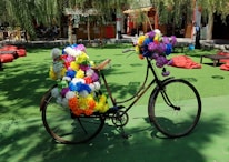 A bicycle is adorned with numerous brightly colored artificial flowers, parked on a green surface in an outdoor area. The setting includes red bean bags scattered around, and a small bar or shop in the background with hanging decorations. Tree branches with leaves are visible above, casting dappled shadows on the ground.