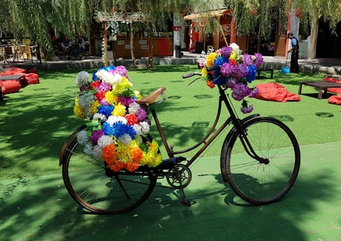 A bicycle is adorned with numerous brightly colored artificial flowers, parked on a green surface in an outdoor area. The setting includes red bean bags scattered around, and a small bar or shop in the background with hanging decorations. Tree branches with leaves are visible above, casting dappled shadows on the ground.