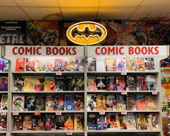 A comic book store display featuring numerous comic books on shelves, with a prominent neon Batman logo at the top. The shelves are labeled with signs reading 'Comic Books' in bright red letters. Various comic book covers with vibrant illustrations are visible, showcasing different characters and themes.