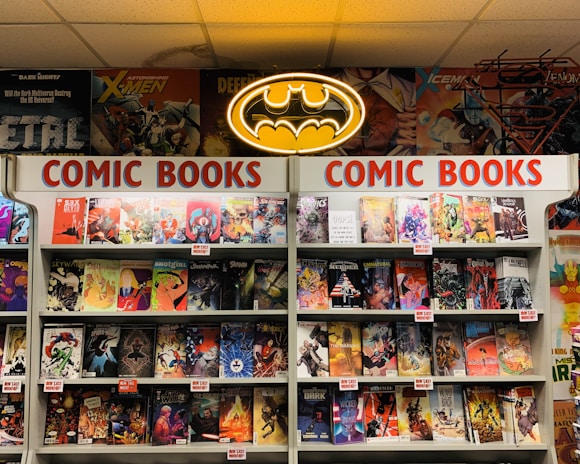 A comic book store display featuring numerous comic books on shelves, with a prominent neon Batman logo at the top. The shelves are labeled with signs reading 'Comic Books' in bright red letters. Various comic book covers with vibrant illustrations are visible, showcasing different characters and themes.