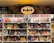 A comic book store display featuring numerous comic books on shelves, with a prominent neon Batman logo at the top. The shelves are labeled with signs reading 'Comic Books' in bright red letters. Various comic book covers with vibrant illustrations are visible, showcasing different characters and themes.