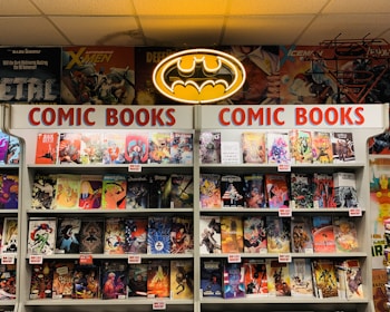 A comic book store display featuring numerous comic books on shelves, with a prominent neon Batman logo at the top. The shelves are labeled with signs reading 'Comic Books' in bright red letters. Various comic book covers with vibrant illustrations are visible, showcasing different characters and themes.