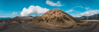 A scenic view of a volcanic landscape with a prominent conical-shaped volcano surrounded by a barren, rugged terrain. The sky above is a clear blue with scattered white clouds. The foreground shows a vast expanse of ash or volcanic sand, while the background features additional mountain ranges.