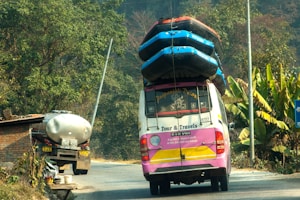A colorful tour van with multiple inflated boats secured on the roof travels on a rural road. Lush green vegetation and banana plants line the side of the road. A tanker truck is parked in front of a brick structure near the road.