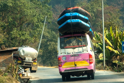 A colorful tour van with multiple inflated boats secured on the roof travels on a rural road. Lush green vegetation and banana plants line the side of the road. A tanker truck is parked in front of a brick structure near the road.