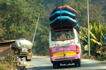 A colorful tour van with multiple inflated boats secured on the roof travels on a rural road. Lush green vegetation and banana plants line the side of the road. A tanker truck is parked in front of a brick structure near the road.