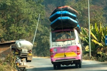 A colorful tour van with multiple inflated boats secured on the roof travels on a rural road. Lush green vegetation and banana plants line the side of the road. A tanker truck is parked in front of a brick structure near the road.