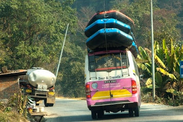 A colorful tour van with multiple inflated boats secured on the roof travels on a rural road. Lush green vegetation and banana plants line the side of the road. A tanker truck is parked in front of a brick structure near the road.