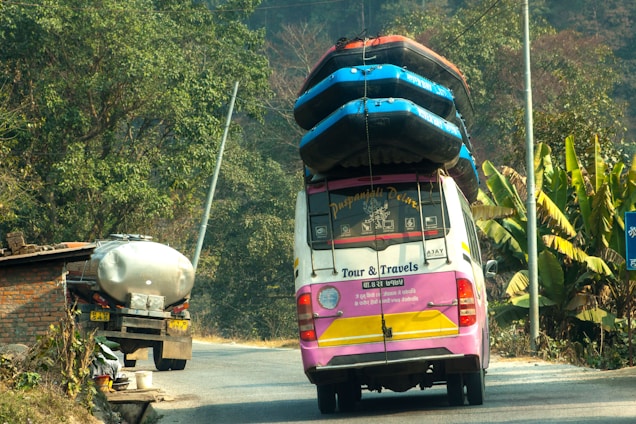 A colorful tour van with multiple inflated boats secured on the roof travels on a rural road. Lush green vegetation and banana plants line the side of the road. A tanker truck is parked in front of a brick structure near the road.