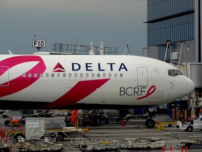 An airplane with Delta Airlines branding is parked at an airport gate. The design features a prominent pink ribbon symbol associated with breast cancer awareness, along with the logo for the Breast Cancer Research Foundation (BCRF). Several ground handling equipment and luggage carts are visible around the aircraft.