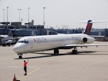 A Delta Air Lines aircraft on the tarmac at an airport, with a ground crew member in a bright orange vest directing it. The background includes an airport terminal and other parked vehicles.