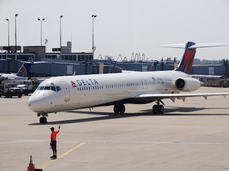 A Delta Air Lines aircraft on the tarmac at an airport, with a ground crew member in a bright orange vest directing it. The background includes an airport terminal and other parked vehicles.