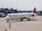 A Delta Air Lines aircraft on the tarmac at an airport, with a ground crew member in a bright orange vest directing it. The background includes an airport terminal and other parked vehicles.