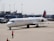 A Delta Air Lines aircraft on the tarmac at an airport, with a ground crew member in a bright orange vest directing it. The background includes an airport terminal and other parked vehicles.