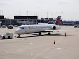 A commercial airplane is parked on an airport tarmac near a terminal. It's a Delta aircraft being guided by a ground crew member in an orange vest. The plane is attached to a tow vehicle at the front, preparing for taxi or maintenance. Various airport equipment and vehicles are present in the background, along with the terminal building.