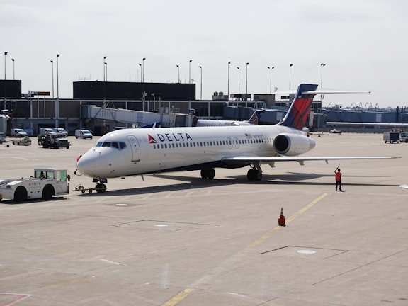 A commercial airplane is parked on an airport tarmac near a terminal. It's a Delta aircraft being guided by a ground crew member in an orange vest. The plane is attached to a tow vehicle at the front, preparing for taxi or maintenance. Various airport equipment and vehicles are present in the background, along with the terminal building.