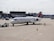A commercial airplane is parked on an airport tarmac near a terminal. It's a Delta aircraft being guided by a ground crew member in an orange vest. The plane is attached to a tow vehicle at the front, preparing for taxi or maintenance. Various airport equipment and vehicles are present in the background, along with the terminal building.