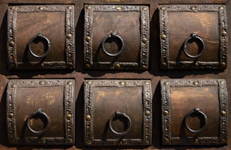 A detailed view of six ornate, square wooden panels, each featuring a decorative metal ring and intricate metalwork along the borders. The wood is dark with visible grain, complemented by the metallic embellishments which include small round studs and decorative patterns.