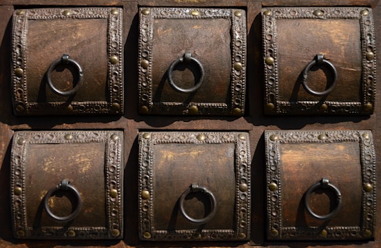 A detailed view of six ornate, square wooden panels, each featuring a decorative metal ring and intricate metalwork along the borders. The wood is dark with visible grain, complemented by the metallic embellishments which include small round studs and decorative patterns.