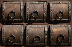 A detailed view of six ornate, square wooden panels, each featuring a decorative metal ring and intricate metalwork along the borders. The wood is dark with visible grain, complemented by the metallic embellishments which include small round studs and decorative patterns.