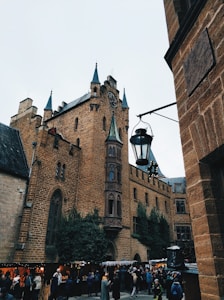 A medieval-style stone building with tall spires and a clock tower, surrounded by greenery. People are gathered below, suggesting a market or festival atmosphere.
