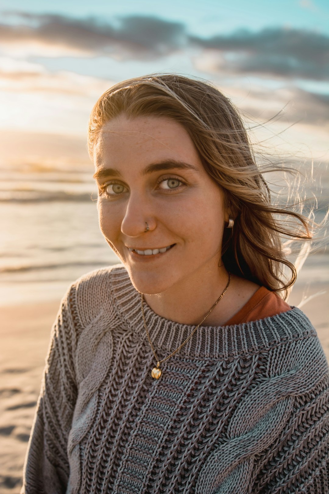 woman in grey knit sweater smiles and stands by the beach