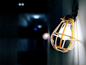 A single light bulb is enclosed in a protective yellow metal cage mounted on a wall, casting a soft glow. The background is dark, creating a stark contrast that emphasizes the illuminated bulb and shadows.