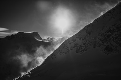 A black and white photograph capturing a dramatic mountain landscape with the sun positioned near the horizon. The light creates a stark contrast, highlighting the rugged textures of snow-covered peaks and atmospheric clouds drifting through the scene.
