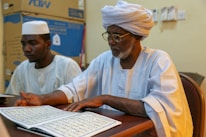 Two men in traditional attire are seated at a table, reading from an open book with Arabic script. One man wears glasses and a white turban, the other a white cap. In the background, there are cardboard boxes.