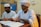 Two men in traditional attire are seated at a table, reading from an open book with Arabic script. One man wears glasses and a white turban, the other a white cap. In the background, there are cardboard boxes.