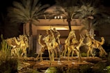 Golden statues of riders on camels are illuminated against a backdrop of tall palm trees and a modern building. The scene is set at night, creating a dramatic and regal atmosphere.