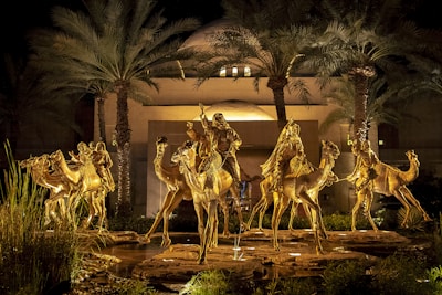 Golden statues of riders on camels are illuminated against a backdrop of tall palm trees and a modern building. The scene is set at night, creating a dramatic and regal atmosphere.