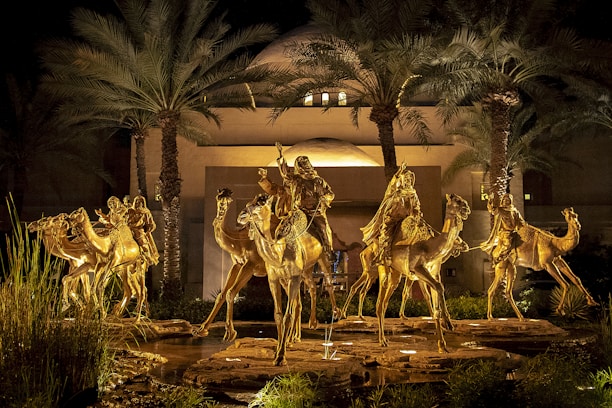 Golden statues of riders on camels are illuminated against a backdrop of tall palm trees and a modern building. The scene is set at night, creating a dramatic and regal atmosphere.