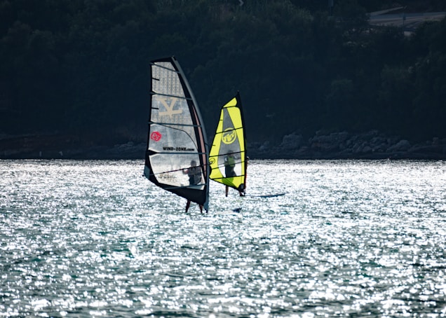 Two windsurfers glide across a sparkling body of water. Their sails catch the light, with one having a dark frame and the other a vibrant yellow color. The water reflects sunlight, creating a shimmering effect, while a shadowy, tree-covered shoreline is visible in the background.