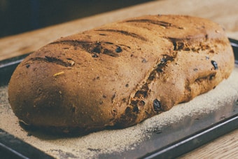 A freshly baked loaf of bread with a golden-brown crust. The bread is placed on a baking tray and appears to have raisins or similar dried fruits embedded in it. The surface of the bread has a rustic texture with several slashes on the top.