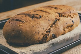 A freshly baked loaf of bread with a golden-brown crust. The bread is placed on a baking tray and appears to have raisins or similar dried fruits embedded in it. The surface of the bread has a rustic texture with several slashes on the top.