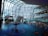 A modern indoor swimming pool area with a large glass ceiling and walls offering a view of snow-covered trees and mountains outside. The pool features a central water fountain, and people are swimming and enjoying the warm indoor environment. A curved bridge spans across the pool, while a staircase and railings are visible on the left side.