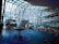 A modern indoor swimming pool area with a large glass ceiling and walls offering a view of snow-covered trees and mountains outside. The pool features a central water fountain, and people are swimming and enjoying the warm indoor environment. A curved bridge spans across the pool, while a staircase and railings are visible on the left side.