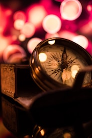 A close-up of a vintage compass perched on a stack of old books with a blurred background displaying circular lights, giving a warm, nostalgic feel.