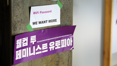 A concrete pillar features two signs taped to it. The top sign is white with the words 'WiFi Password: WE WANT MORE' printed in purple. Below it, a larger purple banner displays Korean text in white, translated as 'Welcome to Feminist Utopia.'