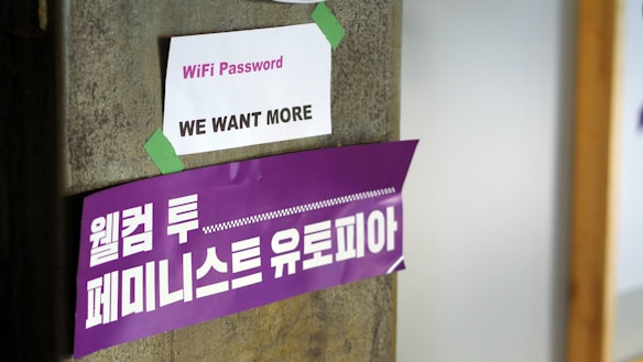 A concrete pillar features two signs taped to it. The top sign is white with the words 'WiFi Password: WE WANT MORE' printed in purple. Below it, a larger purple banner displays Korean text in white, translated as 'Welcome to Feminist Utopia.'