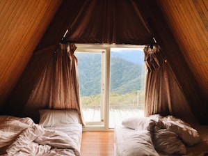 An A-frame cabin room with two unmade beds on either side, positioned under a window with a view of forested mountains. The walls and ceiling are paneled with wood, and the curtains are drawn partially open.