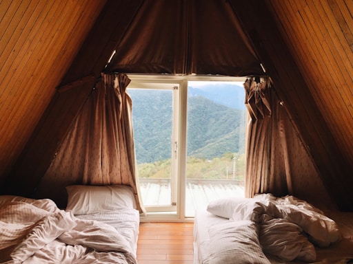 An A-frame cabin room with two unmade beds on either side, positioned under a window with a view of forested mountains. The walls and ceiling are paneled with wood, and the curtains are drawn partially open.
