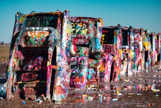 Brightly colored, graffiti-covered cars partially buried with their hoods poking out of the ground, lined up in a row in a muddy field. Spray paint cans are scattered around the base of the vehicles.