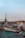 A panoramic view of Parisian rooftops with the Eiffel Tower in the background. The rooftops are characterized by their blue and green hues and visible chimneys. Smoke rises from one of the chimneys, and the sky is overcast with a hint of blue peeking through.