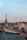 A panoramic view of Parisian rooftops with the Eiffel Tower in the background. The rooftops are characterized by their blue and green hues and visible chimneys. Smoke rises from one of the chimneys, and the sky is overcast with a hint of blue peeking through.